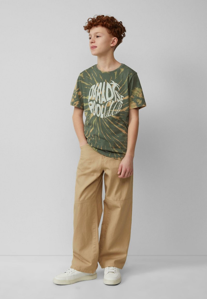 Grünes Batik-T-Shirt mit weißem Aufdruck "PARADISE POOL CLUB", kombiniert mit beigen Schlaghosen und weißen Sneakern.