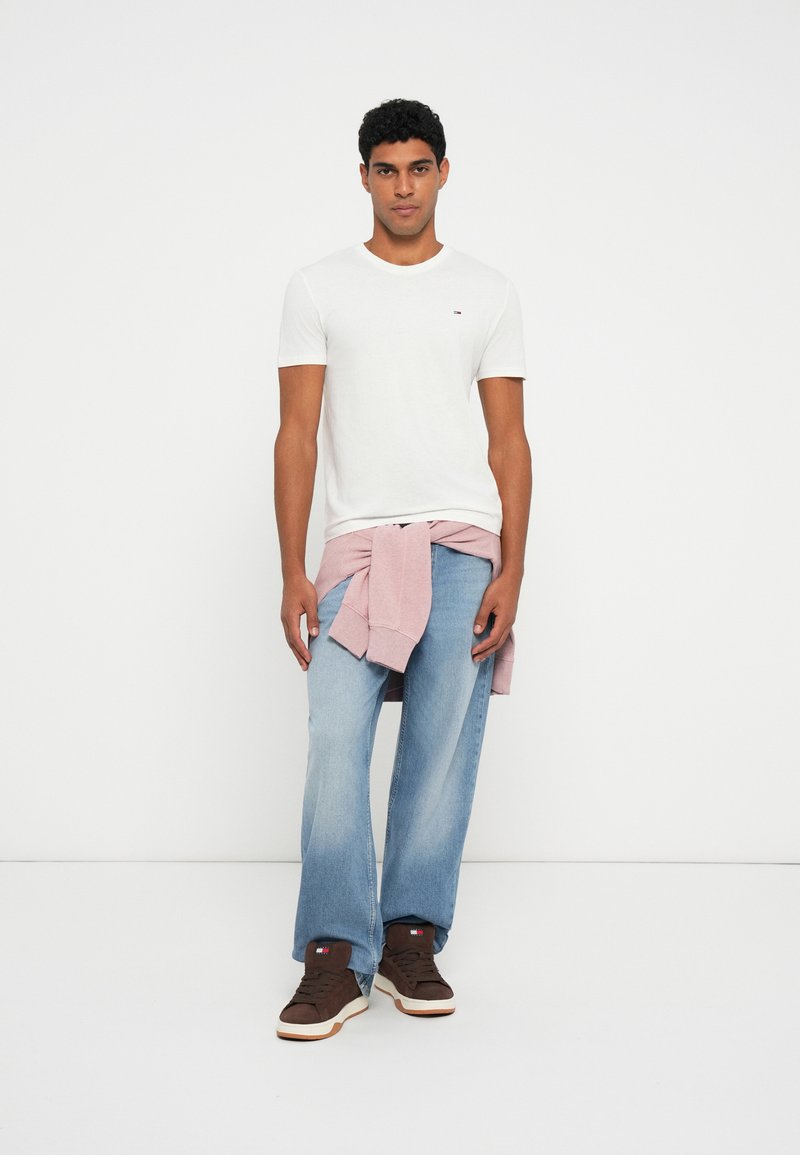 Giovane uomo con una maglietta bianca, jeans azzurri e sneakers marroni, con una felpa rosa legata intorno alla vita, in piedi contro un muro bianco.
