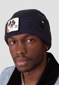 Beanie lavorato a maglia di un blu scuro con una toppa quadrata che presenta un cane ricamato e il testo "TOP DOG". Tessuto morbido, design aderente.
