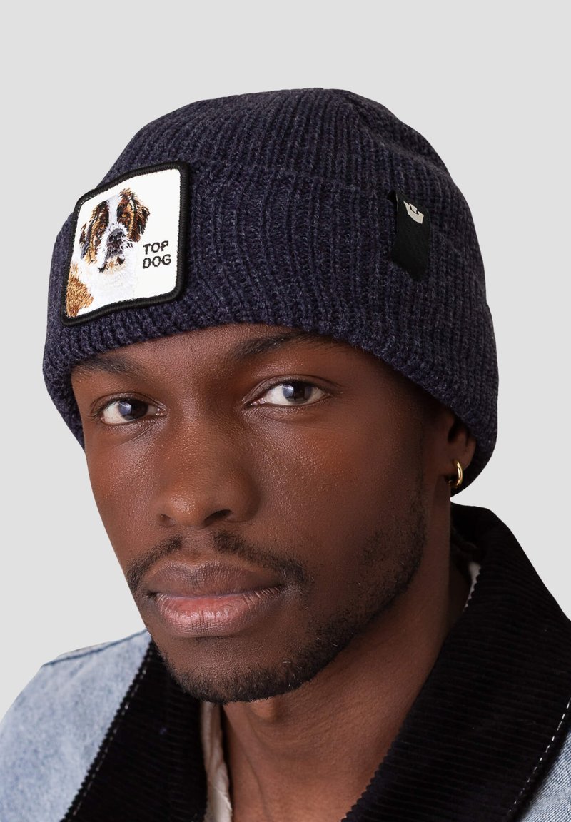 Beanie lavorato a maglia di un blu scuro con una toppa quadrata che presenta un cane ricamato e il testo "TOP DOG". Tessuto morbido, design aderente.