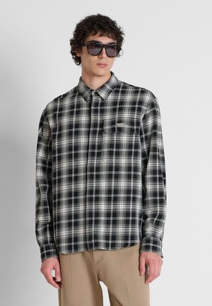 Junger Mann, der ein schwarz-weiß kariertes Langarmhemd, beige Hose und dunkle Sonnenbrille trägt, steht vor einem einfachen hellen Hintergrund.