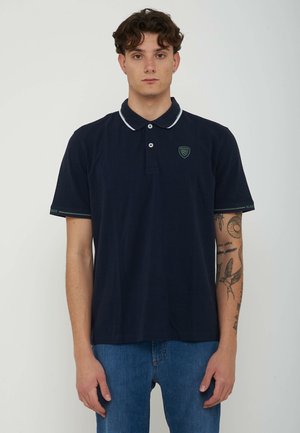 Junger Mann mit kurzen lockigen Haaren, der ein marineblaues Poloshirt und blaue Jeans trägt, steht vor einem schlichten hellen Hintergrund mit sichtbaren Arm-Tattoos.