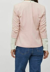 Ljusrosa skräddarsydd kavaj med rynkade ärmar, detalj vid bakseam, ihop med en vit skjorta med knappar under och blå denimbyxor.