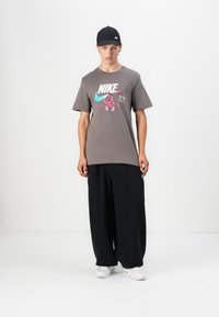 Šedé tričko Nike s vícebarevným grafickým potiskem, s akcenty v odstínech modro-zelené a růžové. V páru s volnými černými kalhotami a bílými teniskami.