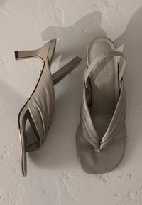 Beige leather sandals with a twisted design, open toe, adjustable slingback strap, and a slim heel. Textured insole detail.
