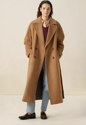 Femme portant un long manteau camel croisé, une chemise rayée, un pull bordeaux, un jean bleu clair et des mocassins à imprimé léopard, adossée à un mur beige.