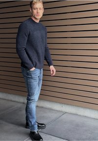 Suéter de punto azul marino con escote redondo, combinado con jeans de mezclilla claros y botas negras hasta el tobillo. De pie contra un fondo de tablones de madera.