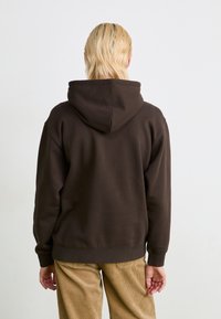 Bruine hoodie met een grote verstelbare capuchon, geribbelde mouwen en zoom. De stof lijkt zacht en dik en wordt gedragen met beige corduroy broek.