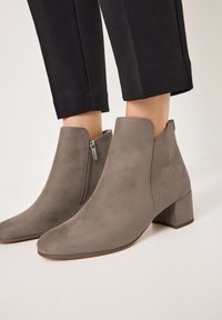 Stivali in suede taupe alti fino alla caviglia con tacco blocco e cerniere laterali, indossati con pantaloni neri accorciati.