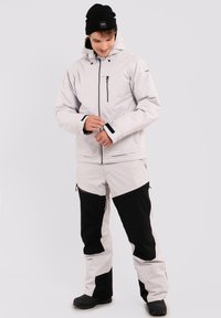 Lichtgrijze jas en broek met zwarte accenten, gemaakt van textuurstof. Voorzien van ritsen en verstelbare manchetten. Zwarte beanie op het hoofd.