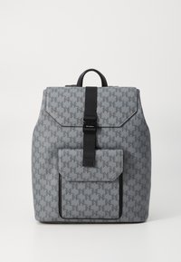 Sac à dos gris à motifs avec un rabat, bandoulière noire et poche avant. Fabriqué en tissu durable, il présente une forme structurée et une surface texturée.