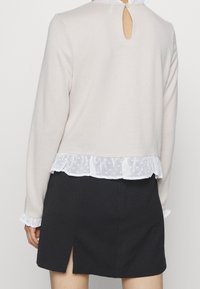 Beiger Pullover mit weißem, gerafftem Spitzenrand und Knopfverschluss am Rücken. Lange Ärmel mit Rüschensaum, kombiniert mit einem schwarzen Rock mit Schlitzen.