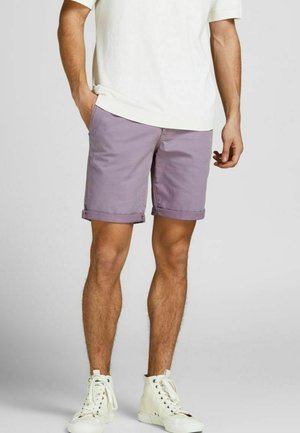 Męskie jasnolawendowe spodenki o klasycznym kroju z wywiniętymi mankietami, wykonane z bawełny. Noszone z białą koszulą z krótkim rękawem i białymi sportowymi butami.