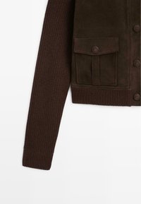 Mørkebrune strikkede ærmer og overholdt suedejakke med knaplukket lommer og store knapper på hvid baggrund.