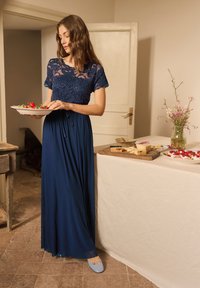 Woman in a navy lace top and long skirt holding a plate of strawberries near a table with cheese and flowers in a simple room.