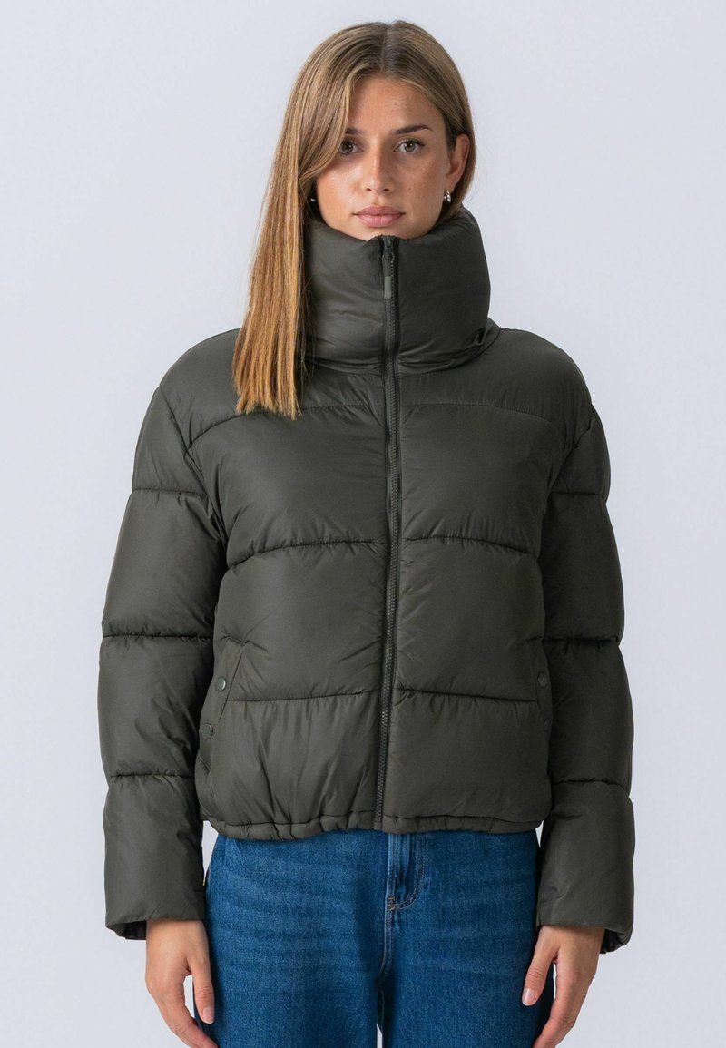 Femme portant une doudoune vert foncé fermée jusqu'en haut, associée à un jean bleu, debout devant un fond uni gris clair.