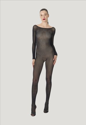 Vrouw met naar achteren gekamd haar, gekleed in een zwart, lang mouwig, geheel lichaam bedekkend netpak en bijpassende open teen hoge hakken, staand tegen een effen achtergrond.