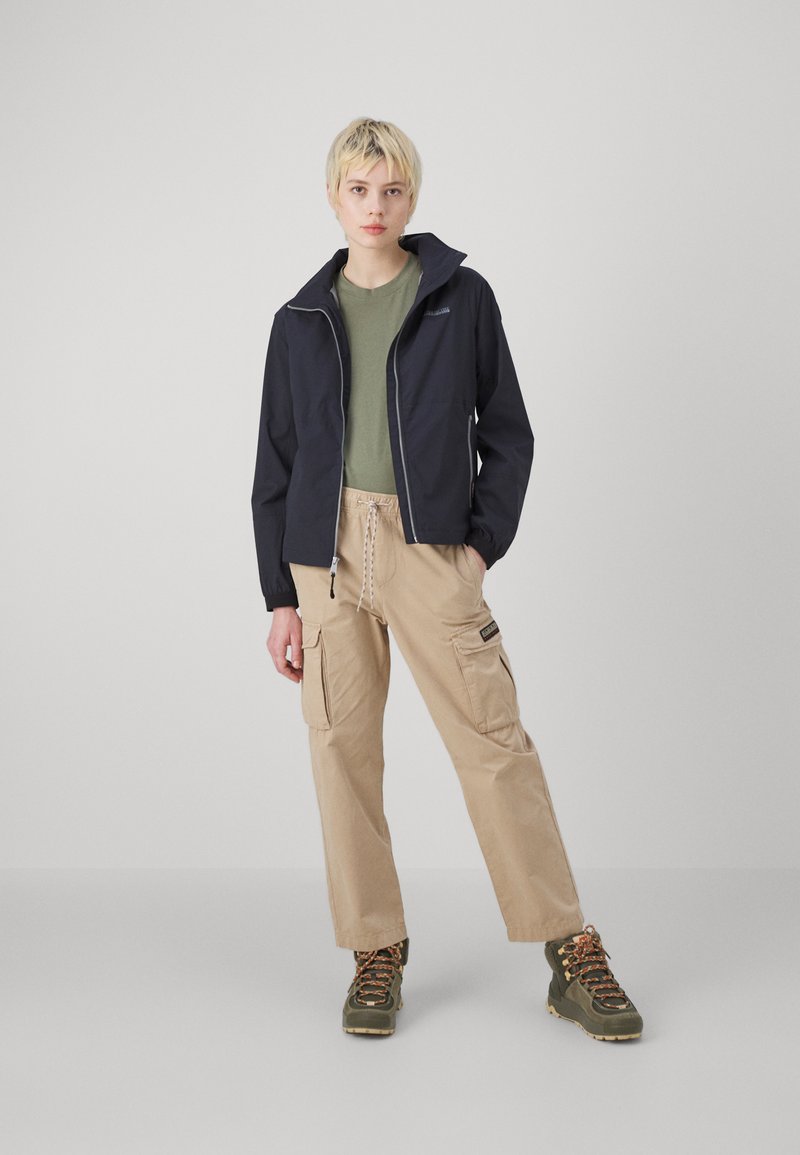 Giacca leggera della marina, camicia verde oliva, pantaloni cargo beige e scarponi da trekking verdi con lacci arancioni, in piedi su uno sfondo bianco.