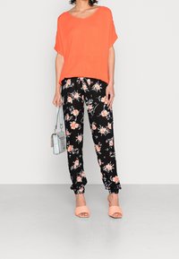 Oranges Kurzarmoberteil, schwarze Hose mit floralem Muster und gekrempelten Knöcheln, pfirsichfarbene Slidesandalen und eine hellblaue Handtasche.