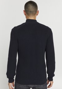 Marineblauer Strickpullover mit hohem Kragen, geripptem Muster, langen Ärmeln und geradem Schnitt. Verfügt über dezente Nahtdetails an den Schultern.