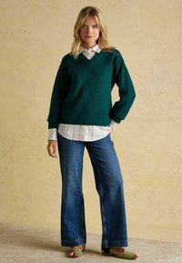 Pulover verde cu decolteu în V, suprapus peste o cămașă albă cu carouri, asociat cu blugi evazați albaștri și pantofi slip-on cu imprimeu. Texturi și culori subtile.