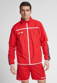 Rød polyester træningsjakke med hvid lynlås og vandret stribe, parret med matchende shorts med sorte chevron-detaljer.