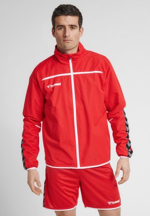 Rød polyester træningsjakke med hvid lynlås og vandret stribe, parret med matchende shorts med sorte chevron-detaljer.