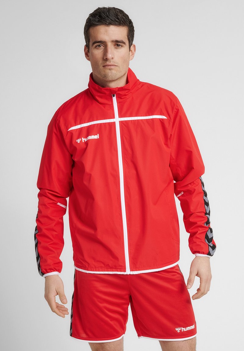 Rød polyester træningsjakke med hvid lynlås og vandret stribe, parret med matchende shorts med sorte chevron-detaljer.