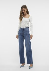 Weißes langärmliges Oberteil kombiniert mit hochtaillierten, weit geschnittenen blauen Jeans und hellblauen Absätzen, was ein lässiges Outfit zeigt.