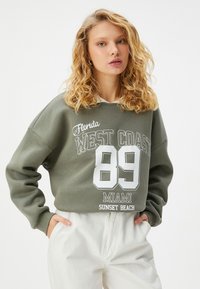 Olijfgroen sweatshirt met witte graphics waarop staat "Florida WEST COAST 89 MIAMI SUNSET BEACH." Heeft een losse pasvorm en geribbelde manchetten.