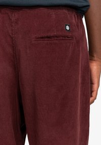 Bordeaux corduroy bukser med vertikal ribbet tekstur, der har en baglomme og en lille logo patch nær taljen.