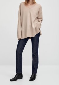 Beige gebreide trui met een V-hals, oversized pasvorm en geribbelde zoom, gecombineerd met donkerblauwe rechte jeans en zwarte enkellaarsjes.