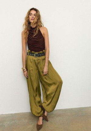 Žena stojící u bílé zdi, oblečená v bezrukávovém hnědém topu, širokých olivově zelených kalhotách stažených černým opaskem a hnědých špičatých botách.