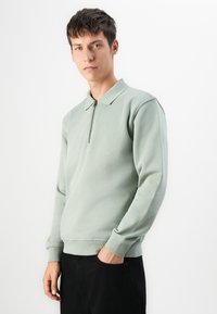 Joven vestido con un pulóver de manga larga verde claro con cuello de cremallera y pantalones negros, de pie contra un fondo blanco.