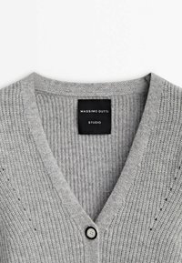 Vaaleanharmaa neulottu neuletakki, jossa on V-pääntie, raihnainen pinta, pieni napitus ja etiketti, jossa lukee "Massimo Dutti Studio."