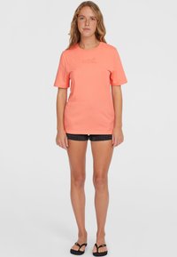 Camiseta de algodón coral con mangas cortas, escote redondo y un sutil detalle del logotipo en un color de contraste. Llevada con pantalones cortos negros y chanclas.