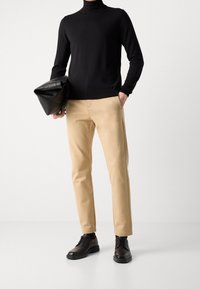 Schwarzer Rollkragenpullover, beiges Hosen und schwarze Lederschuhe. Eine schwarze Leder-Clutch-Tasche in der linken Hand gehalten. Glatte Texturen und taillierte Passform.