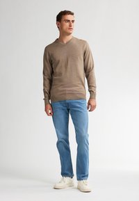 Pulover maro cu decolteu în formă de V, textură tricotată, asociat cu blugi de culoare albastru deschis și adidași albi. Design simplu cu manșete și tiv ribate.
