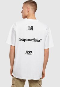 White oversized T-shirt featuring black text: “compton athletics” and “1999” with logo details on the back. Short sleeves, cotton material.