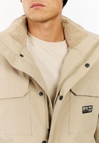 Beige takki, jossa on pehmeä, teksturoitu kaulus, vetoketjusuljenta ja mustat napit. Sisältää rintalogon ja sivutaskut.