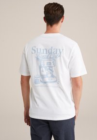 Balta īsām piedurknēm T-kreklu ar zilu grafisko druku, attēlojošu espresso aparātu, un uzrakstu "Sunday Roast Club" aizmugurē.