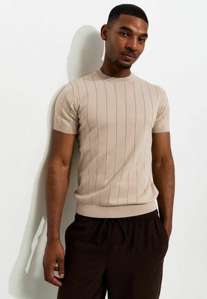 Man met kort haar die een beige kortgebreide trui met ribbelpatroon en donkere broek draagt, staat tegen een witte muur.