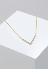 Collana d'oro con un pendente a forma di V adornato di pietre trasparenti, su una catena fine, esposta su un piedistallo grigio chiaro.