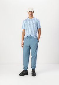 Lichtblauw T-shirt met patroon en korte mouwen, gecombineerd met lichtblauwe elastische broek en zwarte sportschoenen. Pet op het hoofd.