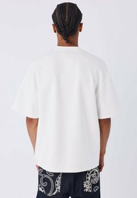 Persona con trenzas que lleva una camiseta blanca oversized y sencilla, y jeans oscuros con patrones bordados en blanco, visto desde atrás.