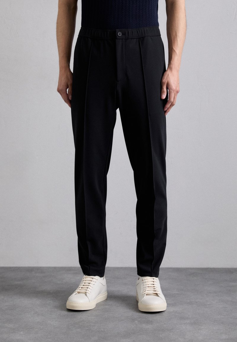 Schwarze maßgeschneiderte Hosen mit einem enganliegenden Bund, ausgestattet mit zwei vorderen Falten und einem glatten, strukturierten Stoff. Kombiniert mit hellen Sneakers.