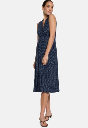 Žena v tmavomodrých midi šatách bez rukávov s bielym bodkovaným vzorom, hnedé sandále na podpätku, stojaca s rukami uvoľnene pozdĺž tela.