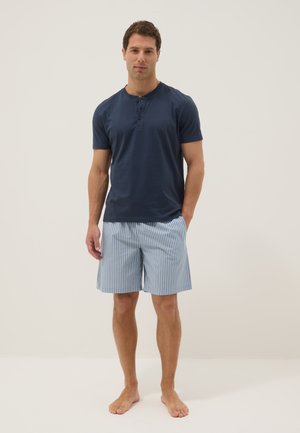 Homme portant un t-shirt henley bleu marine à manches courtes et un short rayé bleu clair et blanc, debout pieds nus sur un fond uni.