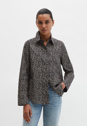 Kobieta ubrana w czarną koszulę na guziki z wzorem, o szerokich rękawach, oraz jasnoniebieskie dżinsy, stojąca na tle jednolitego jasnego tła.