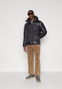 Homme portant une doudoune noire, un pantalon en velours côtelé marron, des chaussures de randonnée et des lunettes de soleil sombres, se tenant devant un fond blanc uni.